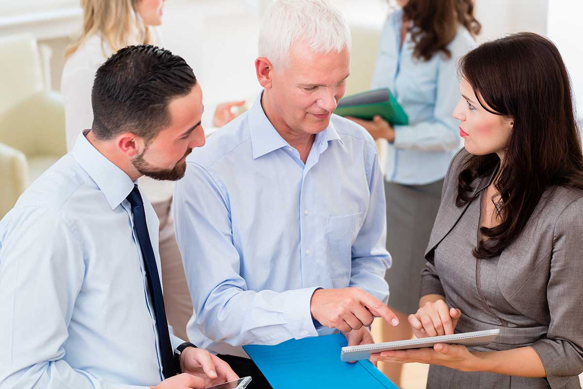 A senior man with white hair points at a tablet held by a woman in a grey dress, while a younger man with a beard looks on, amidst a bright, bustling office background.