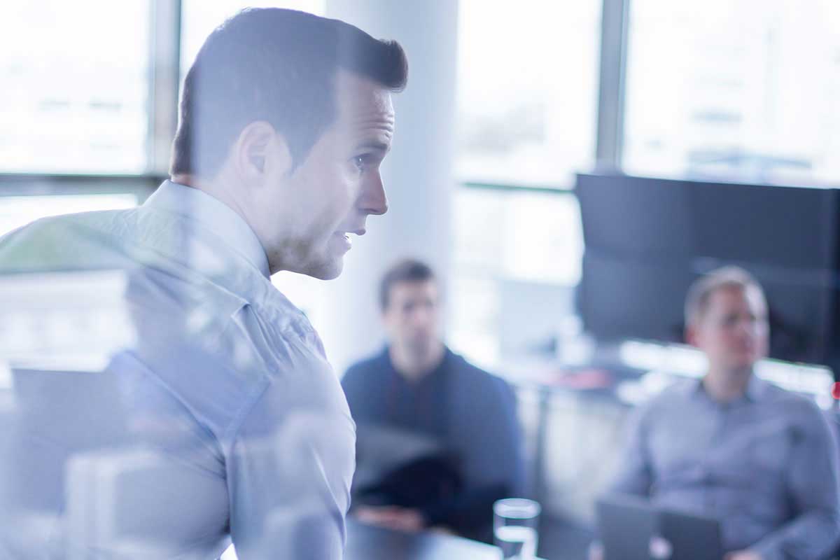 A close-up profile shot of a businessman in a light shirt speaking during a meeting, captured through a glass reflection with two colleagues listening in the blurred background