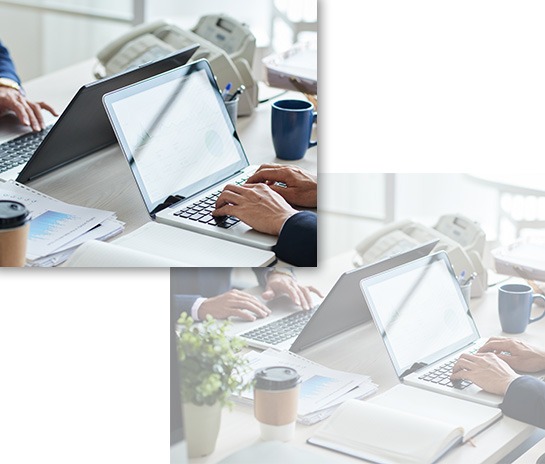 A close-up view of a person's hands typing on a laptop displaying data charts on a white desk, with another person working on a laptop in the background next to coffee cups and printed analytical documents.