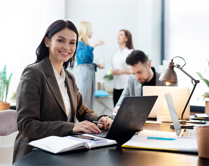 A smiling woman with dark hair wearing a business blazer sits at a desk, typing on a laptop with notebooks and a smartphone in front of her, while colleagues work and talk in a bright, modern office background.